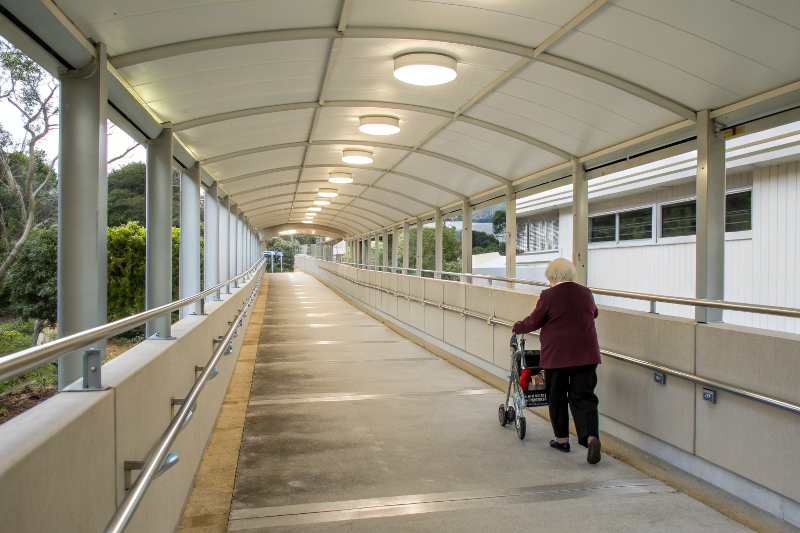 Accessible covered walkway with handrails and LED lighting showing elderly person with walking frame using safe pathway